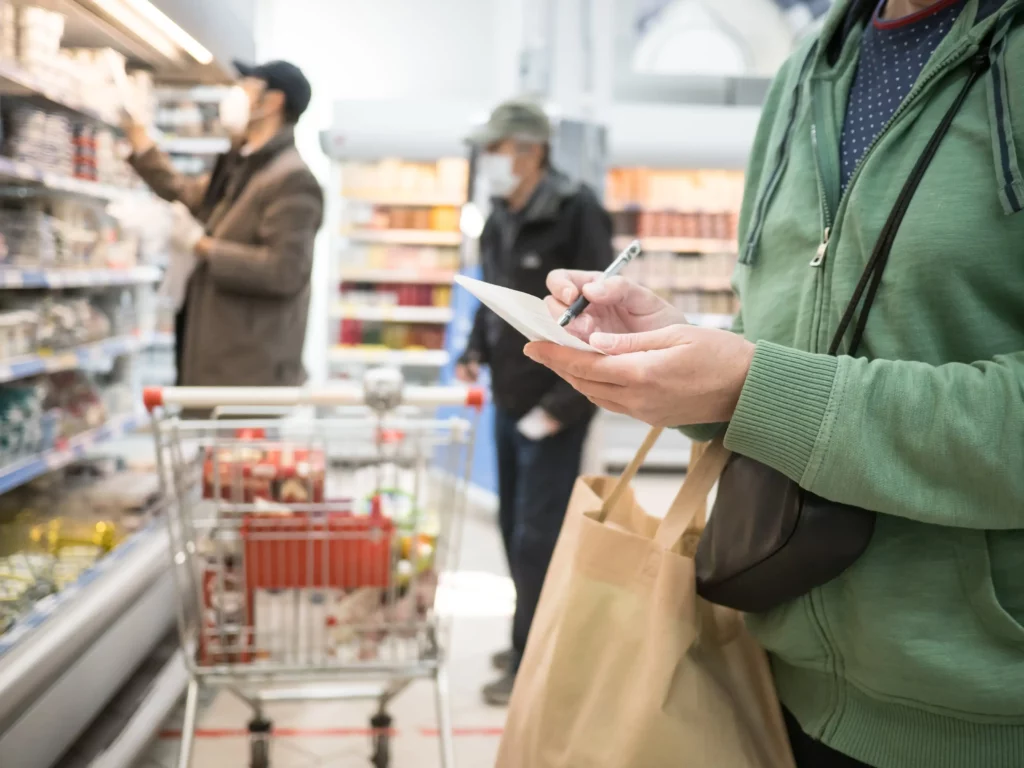 Person mit Einkaufsliste im Supermarkt wählt Grundnahrungsmittel und Notfallbedarf aus